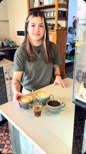 Una chica de Cloe sonriendo detrás de la barra con tres cafés recién hechos.