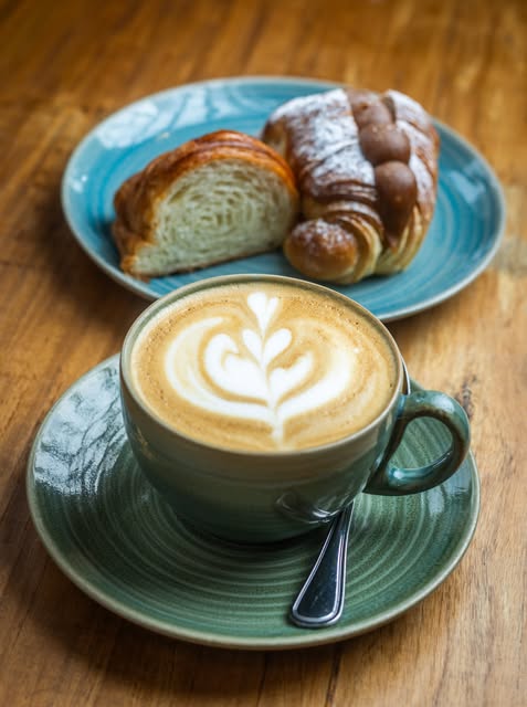 Café con leche con arte latte en taza verde, con un croissant y una medialuna en plato al lado.