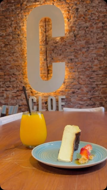 La C de Cloe iluminada en la pared de ladrillo, con un jugo de naranja y una porción de cheesecake en la mesa.